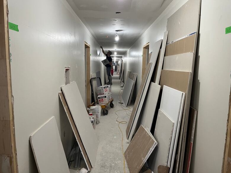 A hallway under construction with white walls and boards scattered around. There is a male worker on a ladder working on the wall in the distance.  