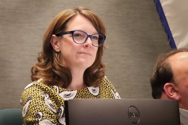 A woman with red hair and glasses wearing a gold print blouse with white birds on it sits in front of a computer screen.