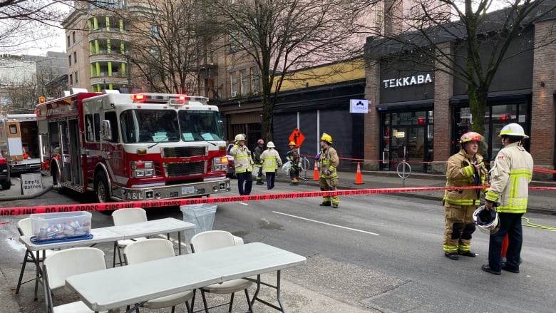 Firefighters converse in front of and behind red caution tape. A fire truck is also visible.