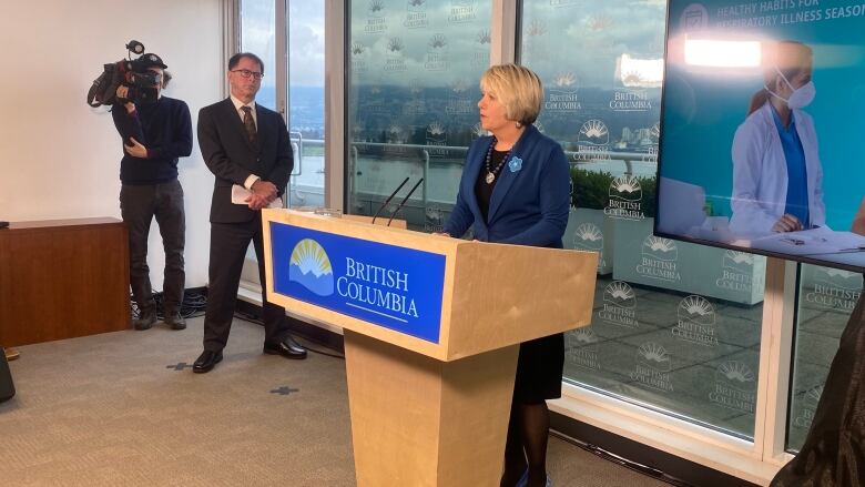 A white woman wearing a dark blue coat speaks at a podium marked British Columbia, with a white man off to her right and a cameraperson behind him.