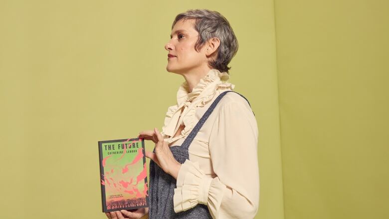 A white woman with short grey hair poses in a side profile with a book. She wears blue overalls and a frilly white shirt. 