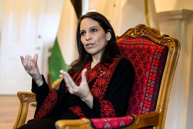 A woman with dark hair sits in a chair and gestures while she speaks. 