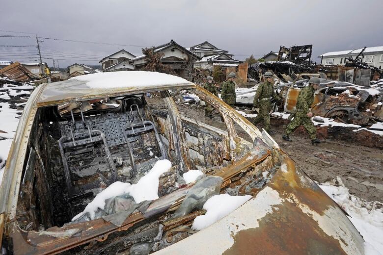 Soldiers in uniform walk along a street where homes are destroyed and lying in rubble under snow. In the foreground is a burned out car.