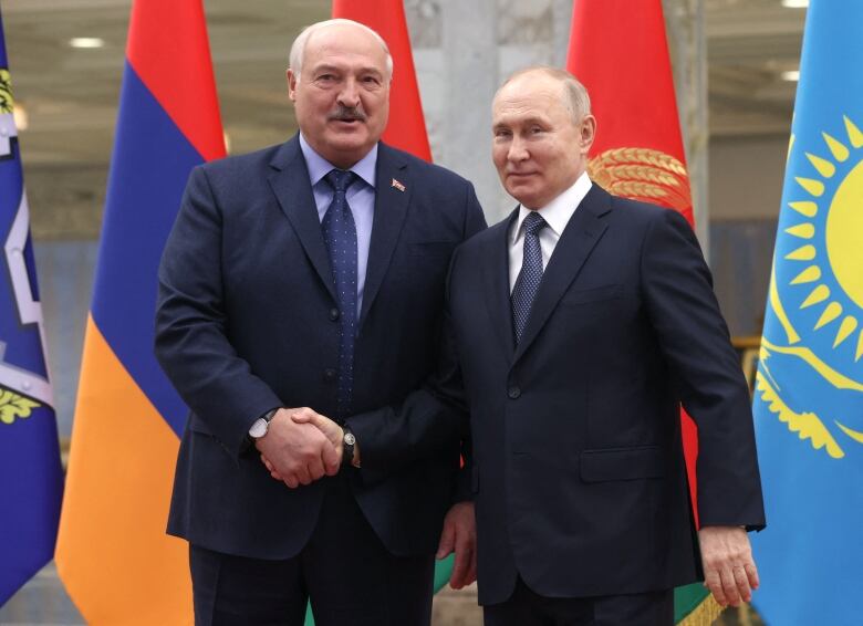 Two men in suits, one mustached, shake hands in front of a number of flags.