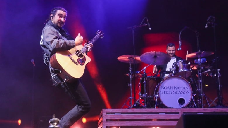 A musician holds a guitar on stage. A drum kit is seen in the background.