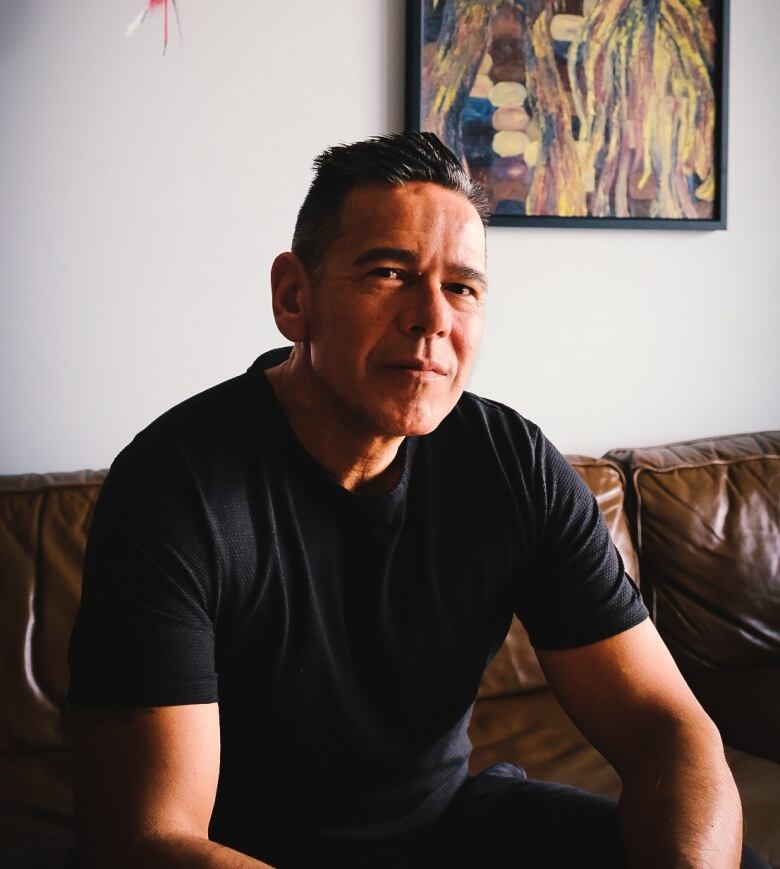 A man wearing a black t-shirt sits on a couch.