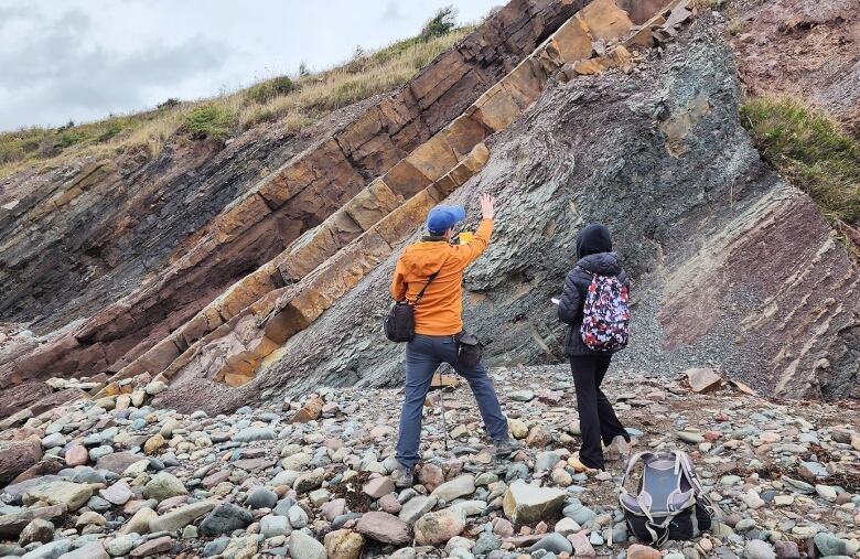 A man in an orange jacket and blue ballcap and a woman in a blue parka with her hood up stand with their backs to the camera, looking up at rock layers.