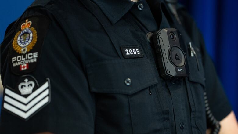 A small camera attached to the front of a button-down black shirt is seen. The shirt's sleeve has a patch with an insignia and text that reads, Police Vancouver. 