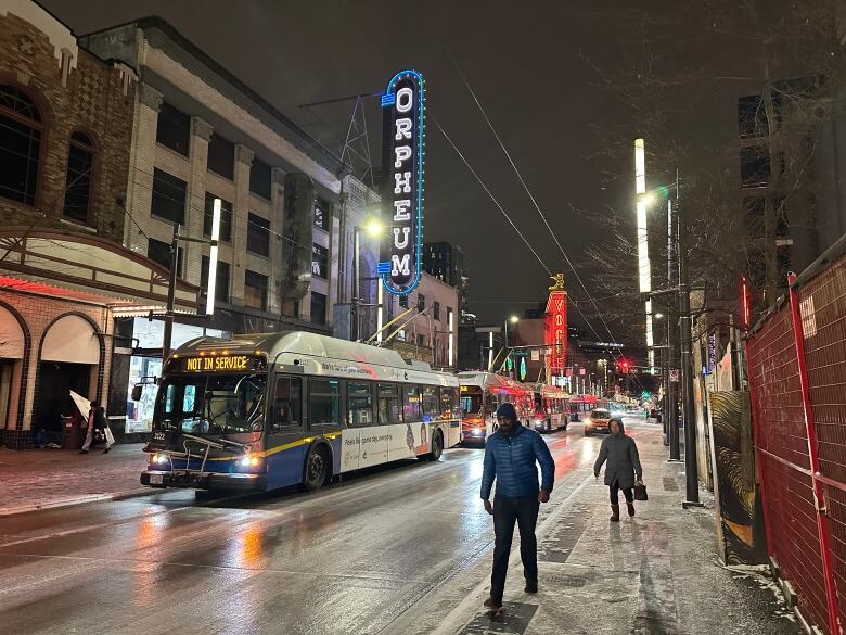 A bus displaying an out of service sign on a dark and slippery road. Pedestrians walk on the sidewalk bent against the cold.