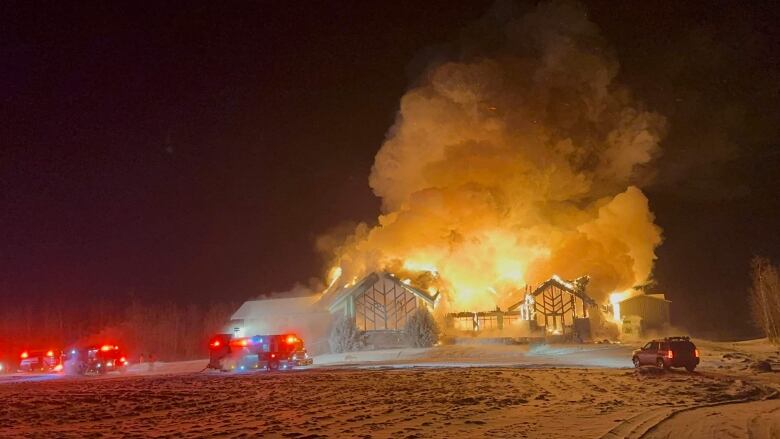 Large clouds of smoke and bright flames stem from a burning golf clubhouse in the middle of the night. Emergency lights flash from three fire trucks parked in front of the building. Tire tracks fill the snow-covered ground in the foreground.