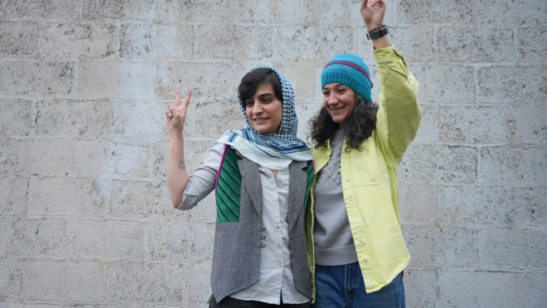 Two women stand together, each raising an arm making peace signs with their hands.