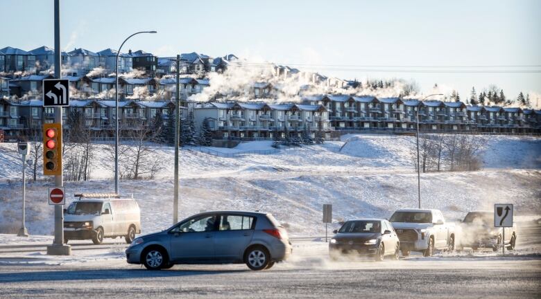 Exhaust from tailpipes of vehicles as they wait at an intersection, with housing up a hill in the background covered in snow.