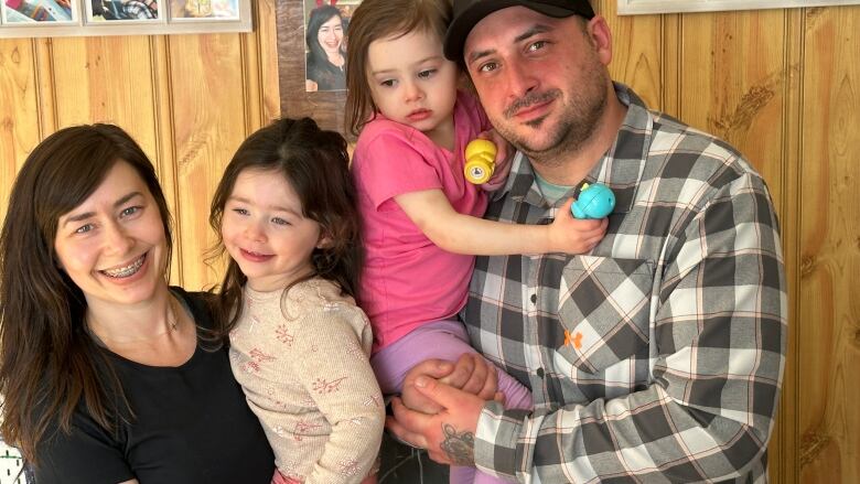 Smiling family. Mom with dark hair, child with dark hair, child with blonde hair and pink shirt and Dad with black baseball cap and black and white plaid shirt stand in front of a pine panelled wall with family pictures.