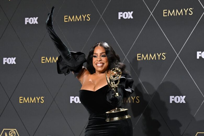 A woman wearing a black dress holds up a golden statuette and raises one arm in the air.