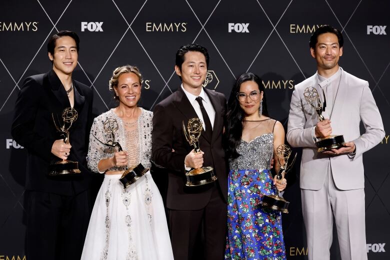 From left, a man in a dark suit, a woman in a shimmery white dress, a man in a black suit, a woman wearing a shimmery silver top and blue skirt, and a man in a gray suit all hold golden statuettes.