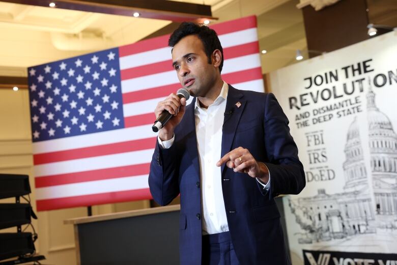 A man wearing a dark suit with a white shirt speaks with a microphone in his hand in front of an American flag and a sign reading 