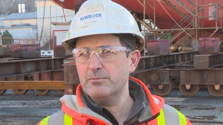 Close up of man wearing eye protection and a white hardhat. Behind him is the hull of a ship.