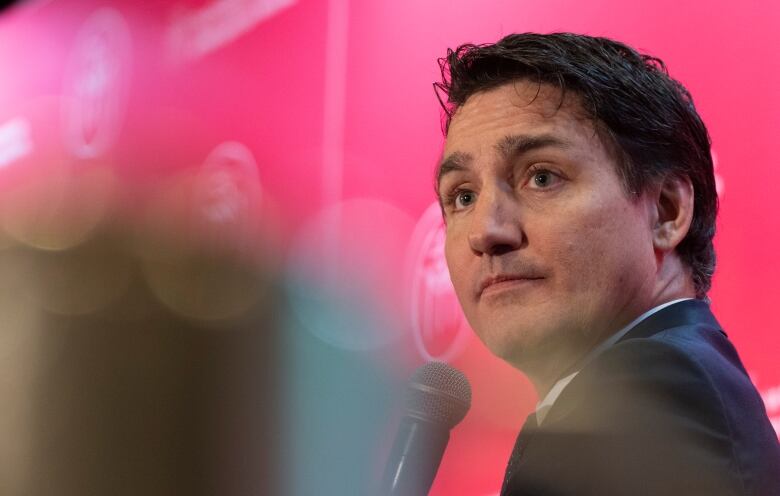 A man in a suit looks into a crowd while holding a microphone. He is sitting in front of a red backdrop.