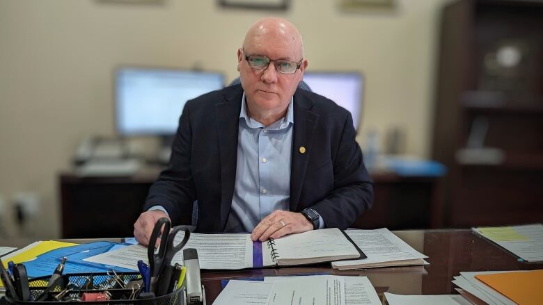 A man in a blue button-up shirt and dark jacket sits at a desk with papers in front of him. 