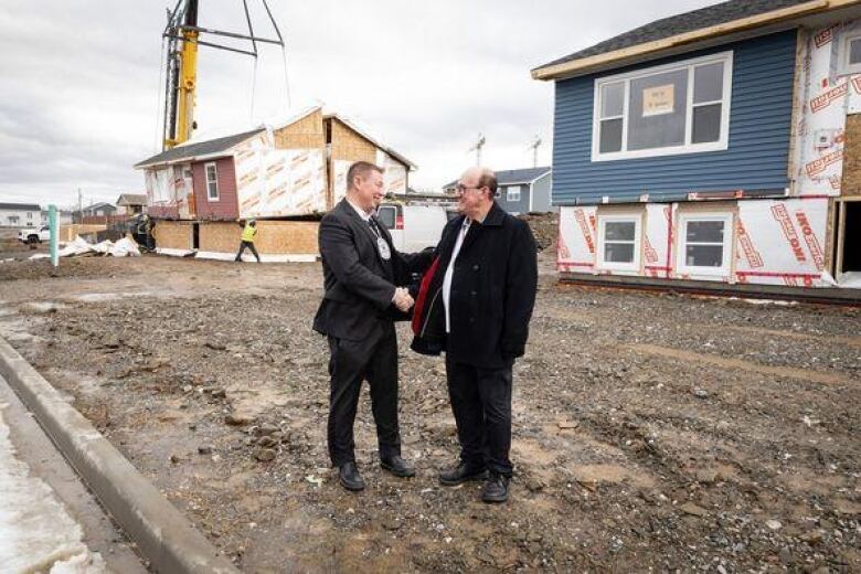 Two men shake hands standing on a dirt yard as construction workers use a crane to lift pieces of a new house into place.