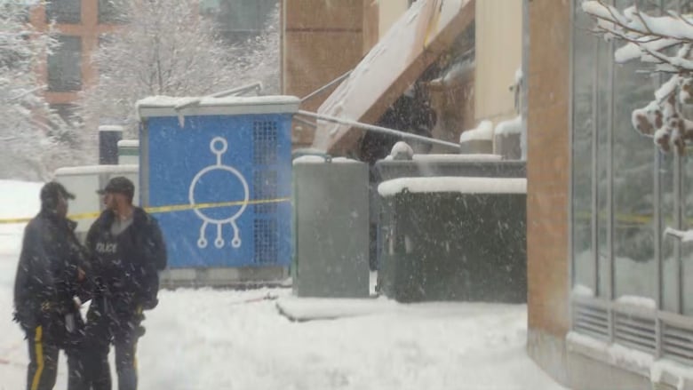 Police officers stand in the snow  in front of a concrete parkade. The shot is fuzzy, but what looks like the shape of a vehicle can be seen sticking out of a hole in the concrete wall.