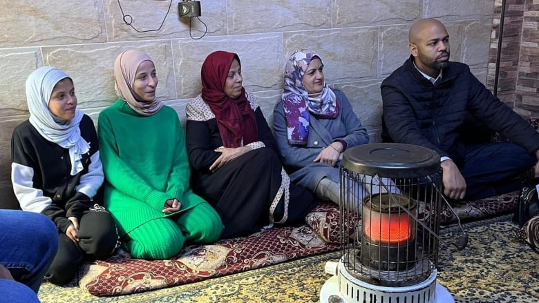 Four women with head scarves and a man on the far right sit on a long cushion. A heating lantern is lit in front of them.