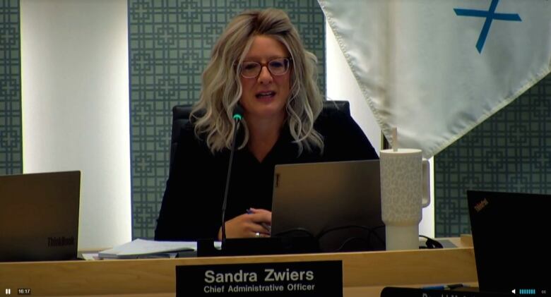 A woman with blonde hair wearing a black suit jacket and glasses sitting in front a laptop and a microphone