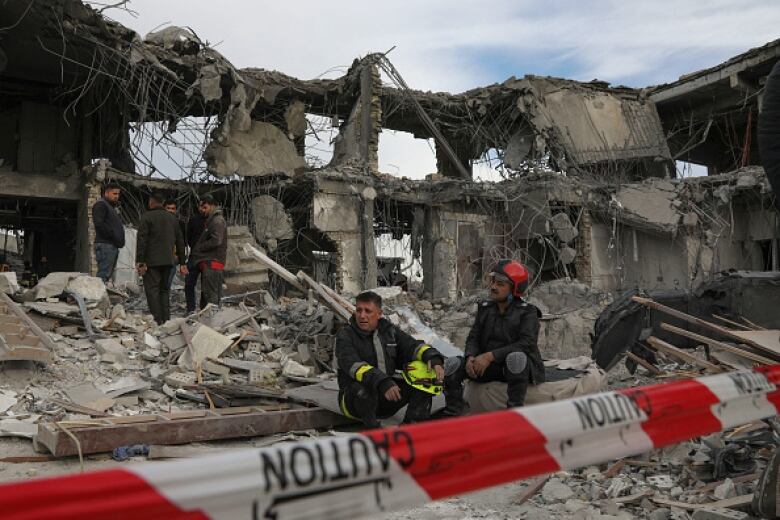 Two men, one wearing a red helmet, sit amid the rubble of a damaged building.