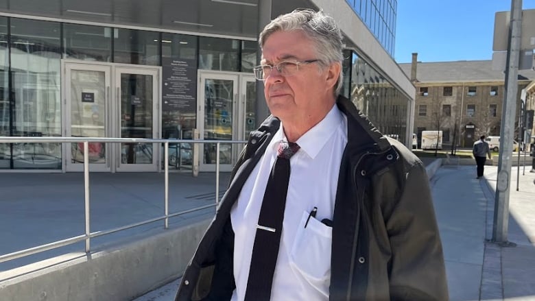 A man with grey hair and glasses walks outside. Behind him is a building with the words Law Courts on it.