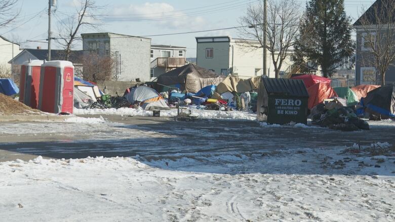 A collection of colourful tents in an open city lot, with two red portable toilet stalls and a metal dumpster. 