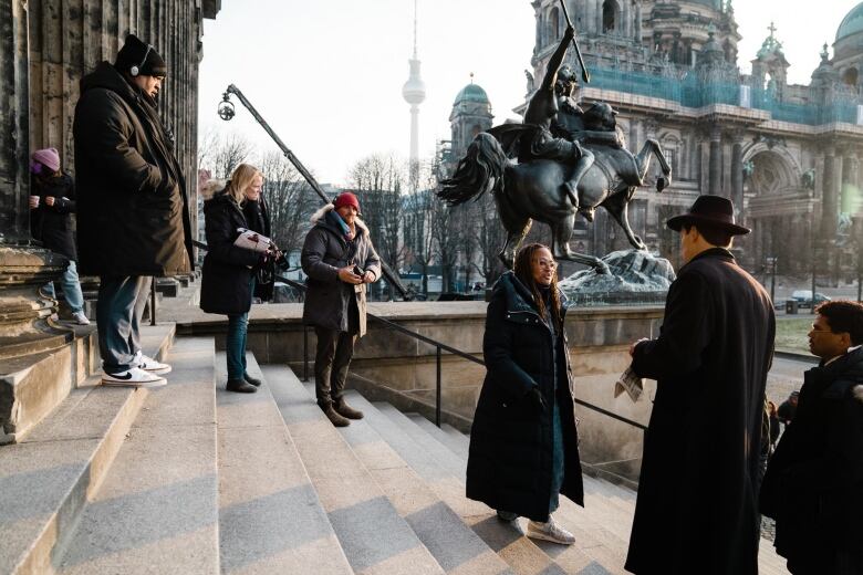 A woman stands on outdoor steps surrounded by actors and film crew with a statue and the Berlin TV tower in the background