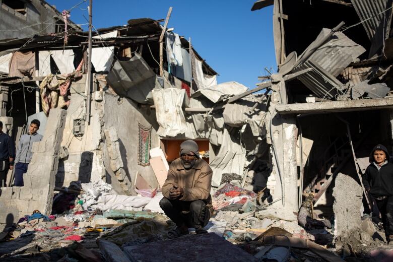 A man crouches on debris in front of bombed homes.