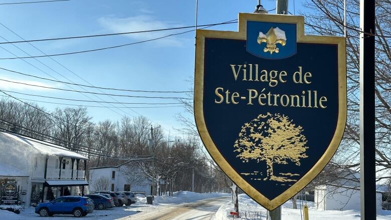 A town sign says Village of Sainte-Ptronille. In behind are houses and a street covered in snow. 