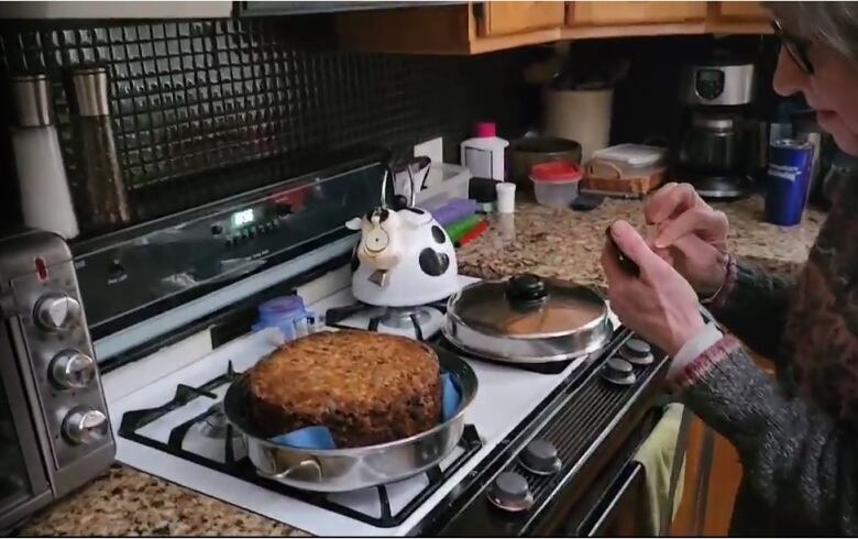 Older woman holding a camera beside a round fruit cake on a white gas stove top.