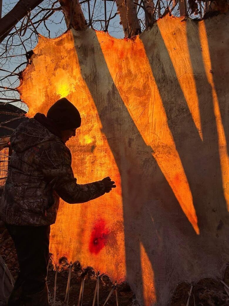 a man in camouflage gear scraping a deer hide at sunset