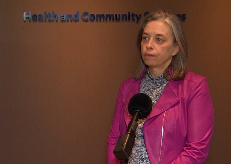 A woman stands against a brown wall and in front of a microphone. She is wearing a bright pink jacket. 