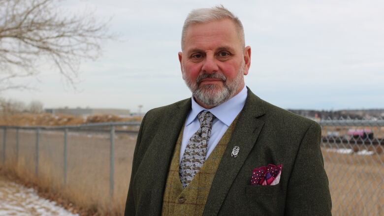 A man with short hair wearing a blue shirt, green vest and green tweed jacket stands in front of the railway tracks in Cochraine, Alta. on an overcast day.