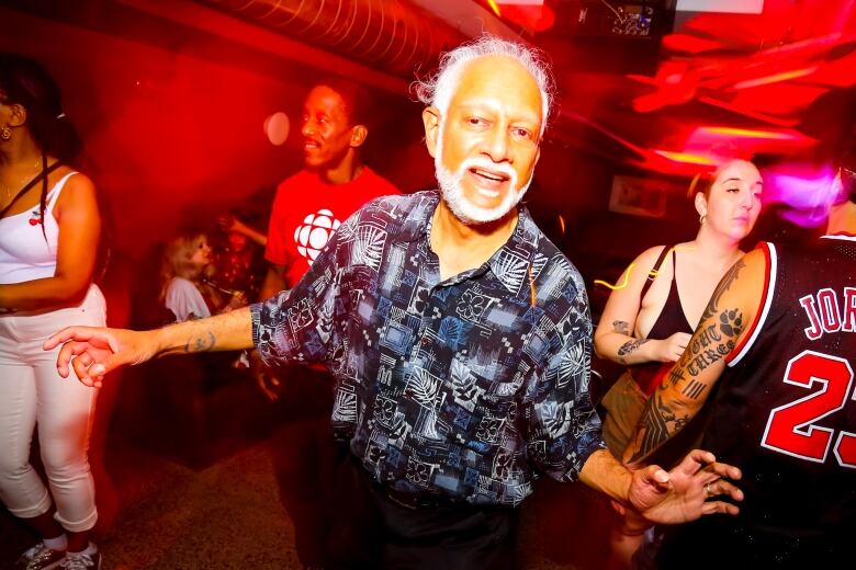 A man in a button-up dances in a club. Several people stand around him. 