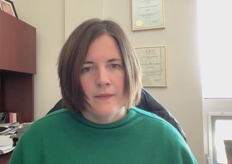 A woman with short brown hair and a green sweater sitting in an office with diplomas in the background.