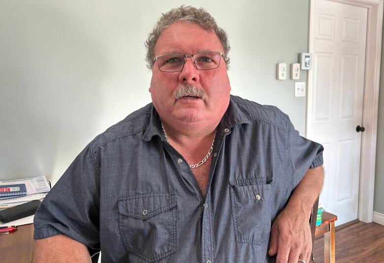 A man with a mustache and glasses, wearing a short-sleeved blue shirt, sits in a room. 