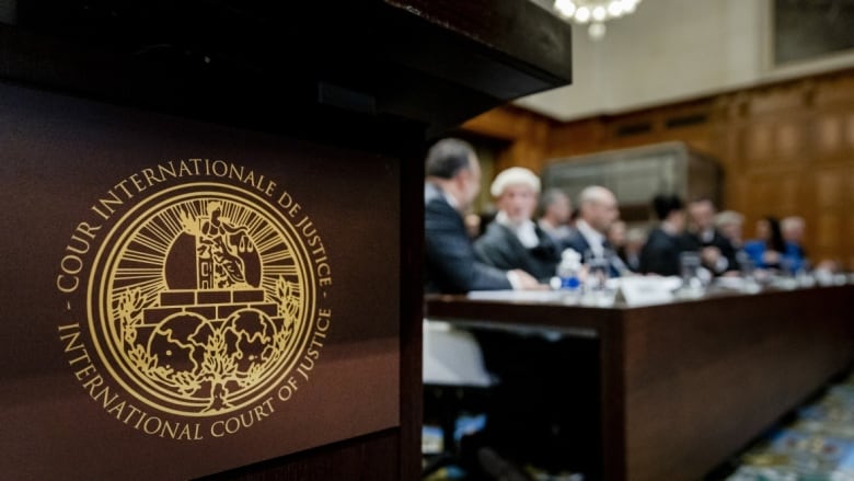 A closeup of a logo reading The International Court of Justice is shown, with the background displaying individuals seated at a table in a courtroom.