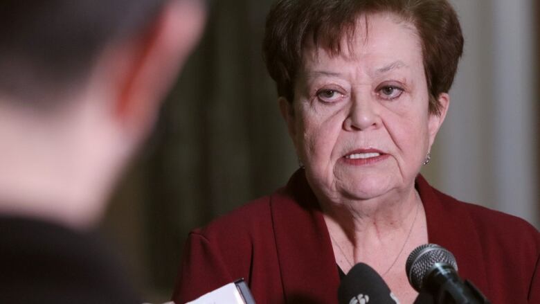 A woman in a red blazer stands in front of several microphones. 