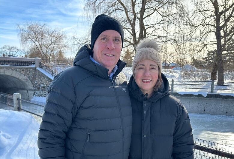 A man stands on the left of a photo. On his right is a woman. The man is wearing a black toque and jacket while the woman is wearing a light brown hat and black jacket.