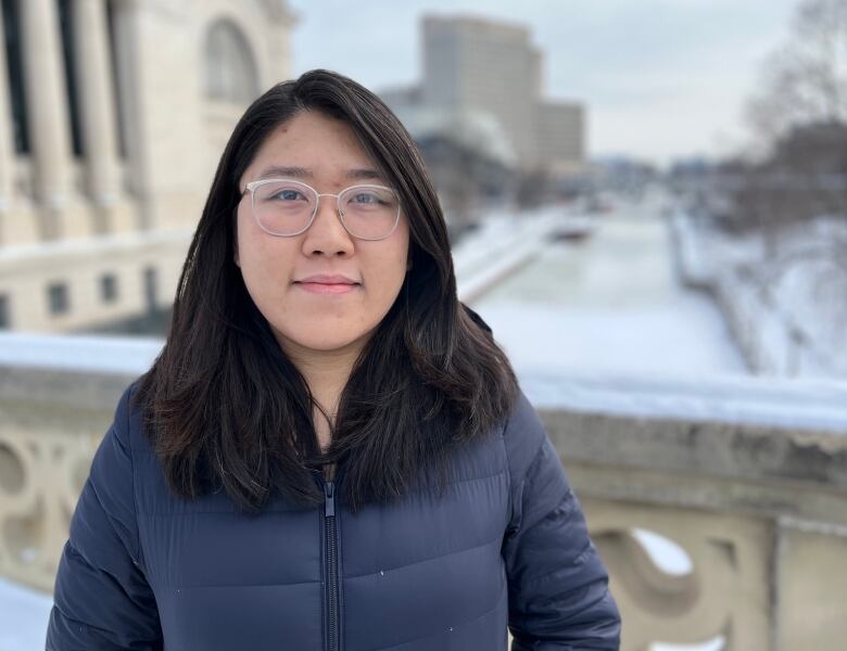 An Asian women with white glasses wearing a blue puffer coat stands on a bridge overlooking the Rideau Canal. She has long black and brown hair.