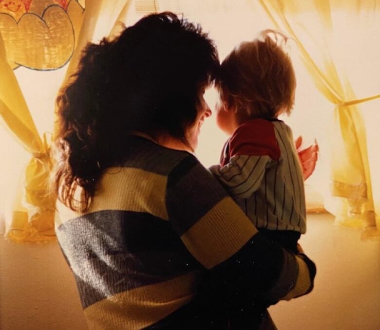 A woman and a young toddler are partially silhouetted as they look out a bright window framed by curtains.