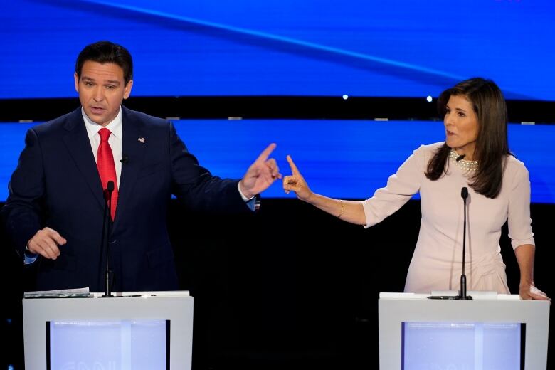 Two people standing at lecterns gesture at each other during a debate.