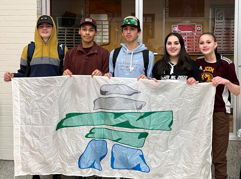 Five young people, three boys and two girls, stand next to each other and smile. They hold up a flag with an inukshuk on it.