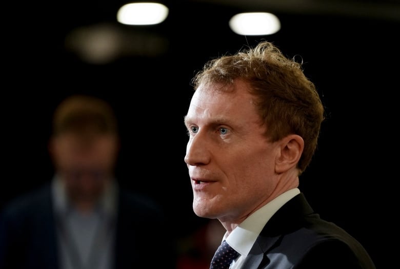 Profile headshot of Immigration Minister Marc Miller, with person standing in the background who is blurred by a shallow depth of field.