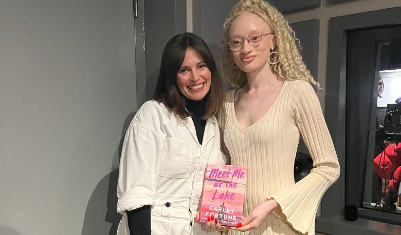 A white woman wearing a white jumpsuit stands next to a Black woman with albinism holding a pink book cover. 