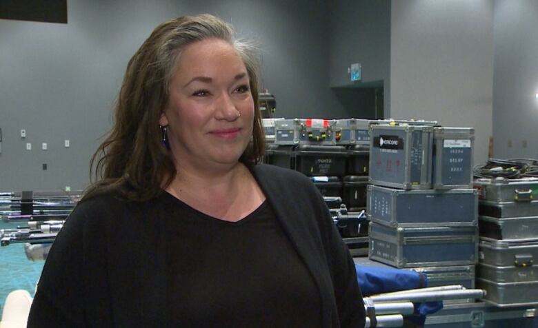  A woman with long, dark hair wearing a black sweater and black shirt looks off to the side of the camera, with metal boxes in the background.